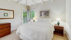 A bedroom with a bed, a dresser, a nightstand with a lamp, a framed picture, and a window with a view of greenery.