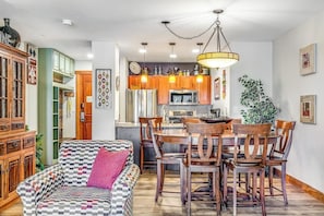 Ein gemütliches Wohnzimmer mit einer gemusterten Couch, einem Holzespeiseesstisch und Stühlen, einer Küchenzeile mit modernen Geräten und einer hängenden Leuchte.