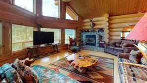 A log cabin interior with a stone fireplace, wooden walls, a television, and a large window.