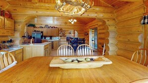 A log cabin interior with a dining table, chairs, a chandelier, and a kitchen area.