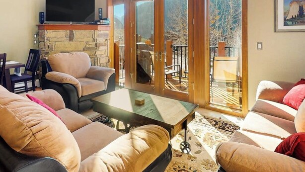 A living room with a stone fireplace, a flat-screen TV, and a large sliding glass door leading to a balcony. - Centrally located with balcony, fireplace, heated garage parking & ski locker (Keystone)