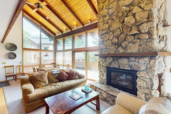 A cozy living room with a stone fireplace, wooden ceiling beams, and large windows. - Conveniently located home with full kitchen, private deck, & loft area (Vail)