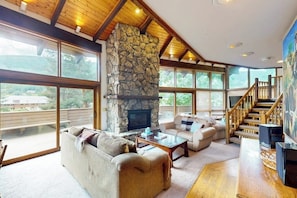 A spacious living room with a stone fireplace, large windows, and a wooden staircase. - Conveniently located home with full kitchen, private deck, & loft area (Vail)