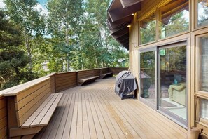 A wooden deck with built-in seating, large glass doors, and a view of a forested area. - Conveniently located home with full kitchen, private deck, & loft area (Vail)