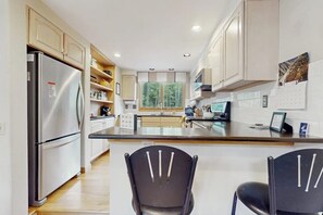 A modern kitchen with a stainless steel refrigerator, wooden cabinets, a central island with chairs, and a window providing natural light. - Charming Vail townhome with private deck that is dog-friendly, bus to slopes (Vail)