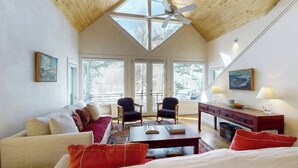 Una habitación con techos altos, techo de madera, un tragaluz grande, un sofá, dos sillones, una mesa de centro, un escritorio y una lámpara.