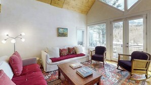 Una sala de estar acogedora con techo de madera, un sofá blanco, un tapete estampado y vistas a un paisaje nevado a través de amplias ventanas.