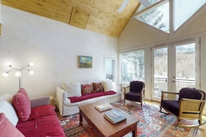 Ein gemütliches Wohnzimmer mit Holzbalkendecke, einem weißen Sofa, einem gemusterten Teppich und Blick auf eine verschneite Landschaft durch große Fenster.