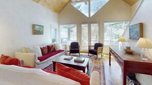 Una habitación amplia con techo inclinado, ventanas grandes, ventilador de techo, un sofá, sillones, una mesa de centro y un escritorio con lámpara.