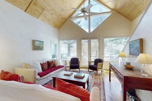 Ein geräumiger Raum mit schrägverdeckter Decke, großen Fenstern, einem Deckenventilator, einer Couch, Sesseln, einem Couchtisch und einem Schreibtisch mit Lampe.