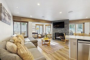 Una sala de estar moderna con un sofá, una chimenea, un televisor montado en la pared y una zona de cocina con lavavajillas.