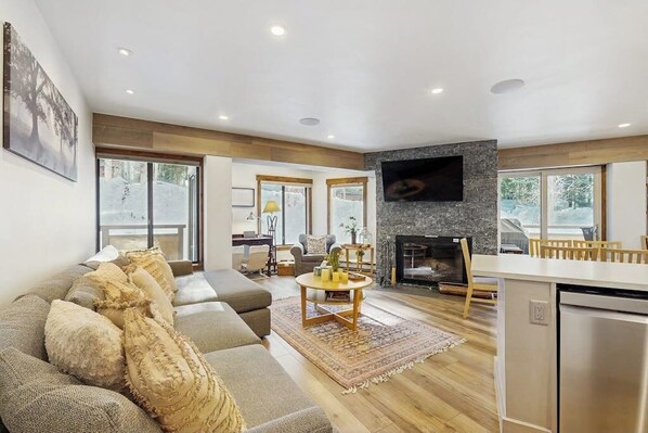 A modern living room with a sofa, a fireplace, a TV mounted on the wall, and a kitchen area with a dishwasher. - Charming condo w/ full kitchen, shared pool & hot tub - near ski resort (Vail)