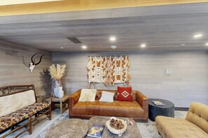 A modern living room with a leather sofa, patterned cushions, a coffee table with books and a bowl, and a decorative wall hanging. - Conveniently located condo w/ fast wifi, shared hot tub & amazing location (Vail)