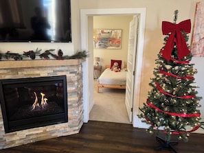 Ein Wohnzimmer mit Kamin, einem darüber montierten Fernseher, einem Weihnachtsbaum mit roter Schleife und einem Bett im Hintergrund.
