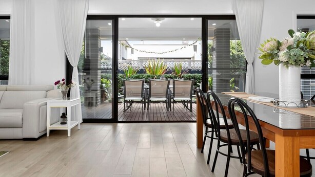 A modern living room with a sofa, a dining table with chairs, a small side table, and a large sliding glass door leading to an outdoor patio with a table and chairs.