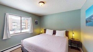 Um quarto com uma cama, duas luminárias de cabeceira, uma janela com cortinas, um quadro na parede e uma luminária de teto.