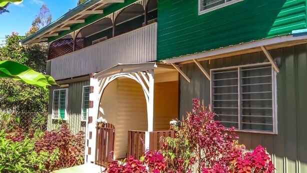 A two-story house with a green roof, a covered porch, and a fenced yard with flowering plants. - Shalom Mission Home (Mount Hagen)