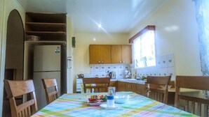 A kitchen with wooden cabinets, a refrigerator, a dining table with chairs, and a window with curtains. - Shalom Mission Home (Mount Hagen)