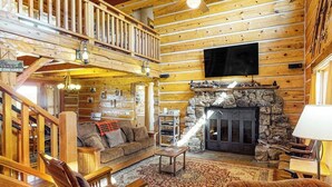 A log cabin interior with a stone fireplace, wooden furniture, a TV mounted on the wall, and a balcony. - Ridgetop mountain-view home on 5 acres with Gas Grill, AC & fireplace (Pagosa Springs)