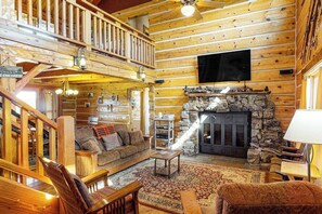 A log cabin interior with a stone fireplace, wooden furniture, a TV mounted on the wall, and a balcony. - Ridgetop mountain-view home on 5 acres with Gas Grill, AC & fireplace (Pagosa Springs)