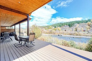 Eine überdachte Terrasse im Freien mit Tisch und Stühlen, mit Blick auf einen Fluss und bewaldete Hügel.