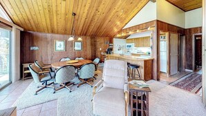 Un comedor con paneles de madera, una mesa grande y sillas, una zona de cocina con mostrador y electrodomésticos, y una puerta que da a otra habitación.