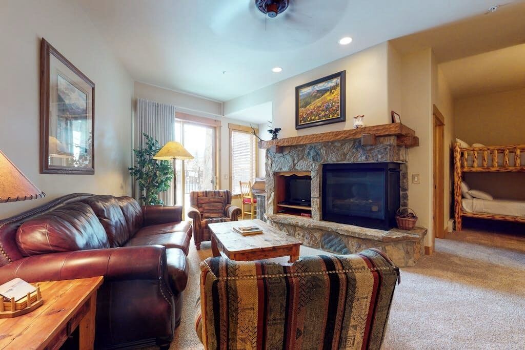 A living room with a leather sofa, a patterned armchair, a wooden coffee table, a stone fireplace, and a television.