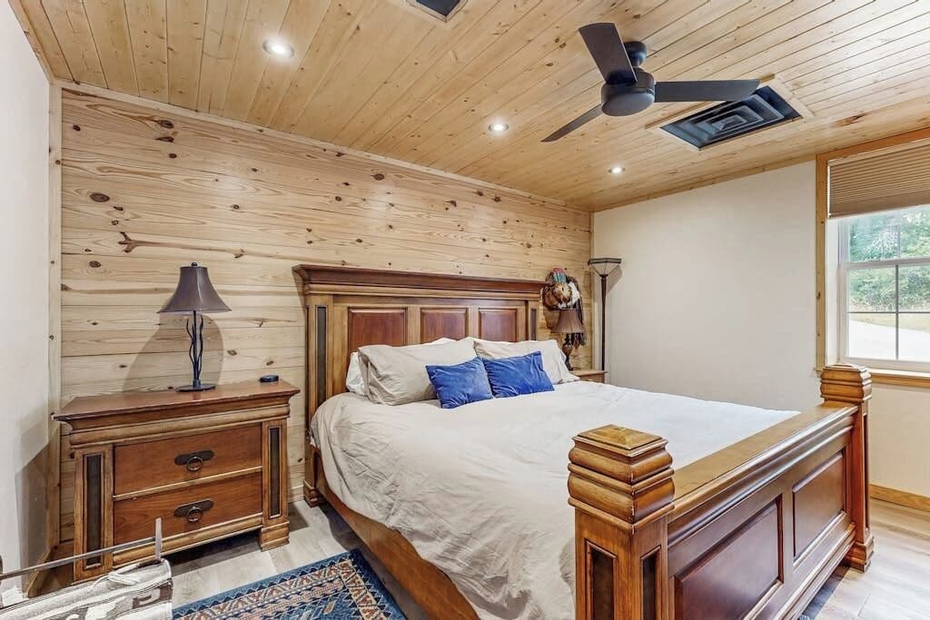 A bedroom with a wooden ceiling, a bed with a headboard, a nightstand, a lamp, and a window.