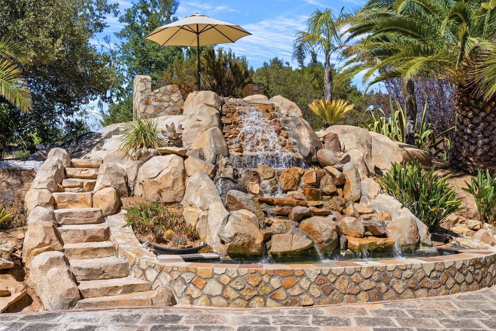 A rock waterfall with a stone staircase leading up to it, surrounded by tropical plants and a palm tree.