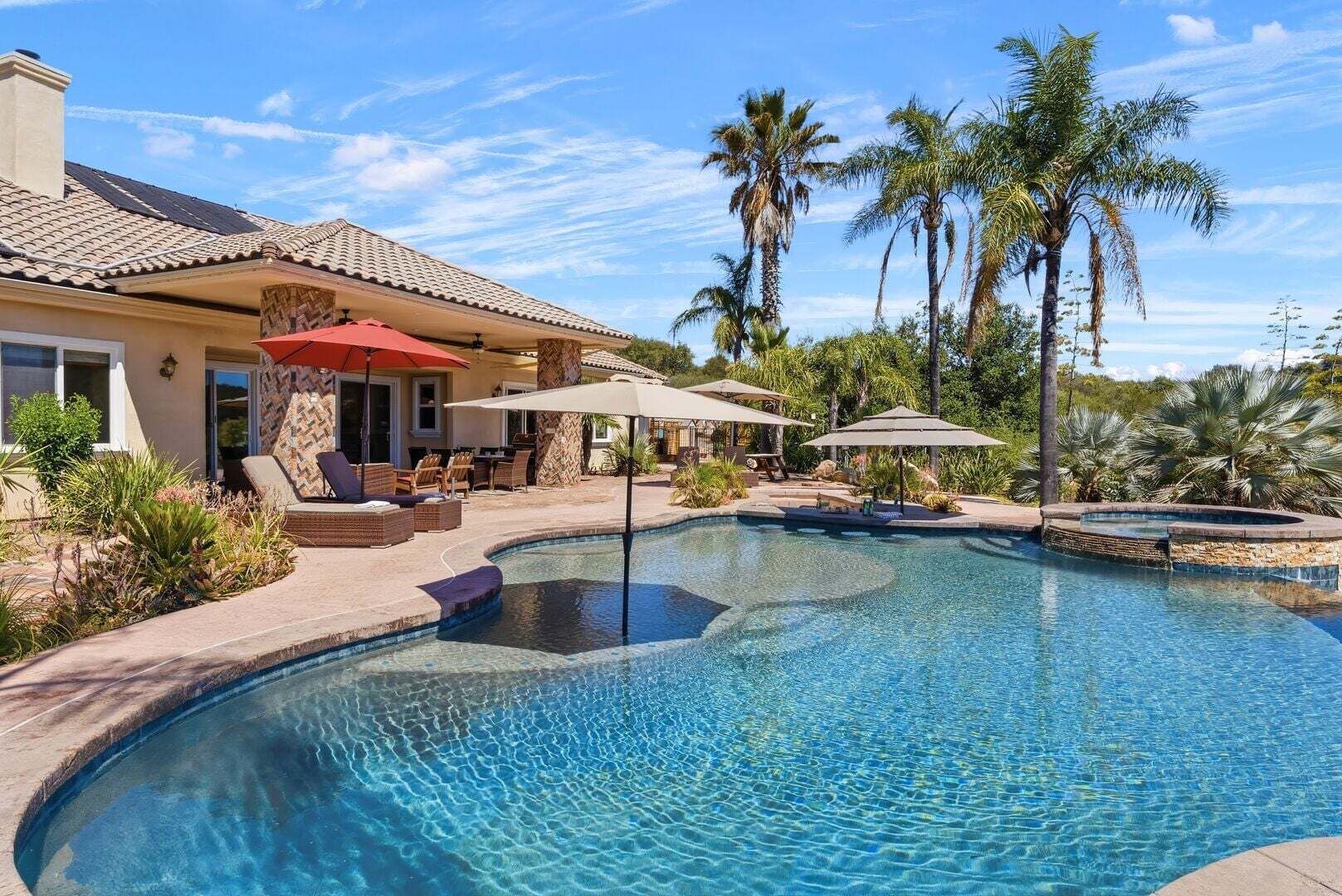 A swimming pool with lounge chairs and umbrellas, surrounded by palm trees and a house.