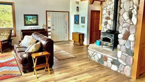 A living room with a stone fireplace, a brown leather sofa, wooden flooring, and a door leading to another room. - Dog-friendly cabin with large yard, wood stove, grill & mountain views (Pagosa Springs)