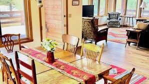 A dining area with a wooden table, chairs, and a TV in the background. - Dog-friendly cabin with large yard, wood stove, grill & mountain views (Pagosa Springs)