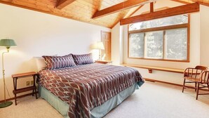 A bedroom with a wooden ceiling, a bed with patterned bedding, bedside lamps, a window with blinds, and a chair. - Dog-friendly cabin with large yard, wood stove, grill & mountain views (Pagosa Springs)