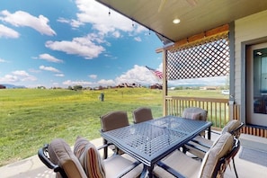 A covered patio with a table and chairs, an American flag, and a view of a grassy field and mountains. - Dog-friendly house in central locale on golf course with mountain view & hot tub (Pagosa Springs)