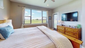 Une chambre à coucher avec un lit, un ventilateur de plafond, une télévision, une fenêtre donnant sur un beau paysage et une commode en bois.