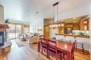 A spacious living area with a fireplace, wooden dining table, and a kitchen with wooden cabinets and a hanging light fixture. - Ground-floor, walk-to-lift condo w/ fireplace, W/D & shared pool/hot tubs/gym (Steamboat Springs)