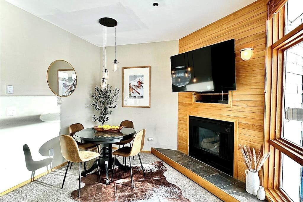 A modern dining area with a round table, chairs, a fireplace, a TV mounted on the wall, and a large window.