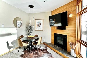 Ein moderner Essbereich mit rundem Tisch, Stühlen, einem Kamin, einem an der Wand befestigten Fernseher und einem großen Fenster.
