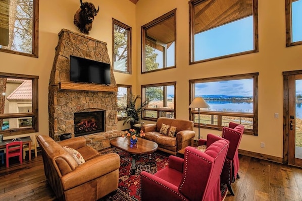 A spacious living room with a stone fireplace, leather sofas, and large windows overlooking a body of water. - Lakefront home with private dock, mtn & lake views, bikes, fireplace, grill, SUP (Pagosa Springs)
