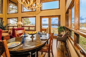 A dining area with a round table, chairs, a chandelier, and large windows overlooking a body of water. - Lakefront home with private dock, mtn & lake views, bikes, fireplace, grill, SUP (Pagosa Springs)