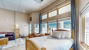 Ein Schlafzimmer mit einem großen Bett, einem Holz-Kopfteil, einem Nachttisch aus Holz, einer Holzkommode, einem braunen Ledersofa, einem blauen Sessel und Blick auf Berge durch große Fenster.