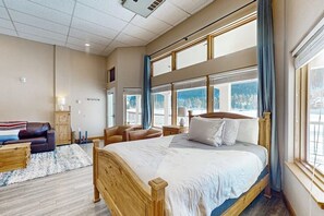 Ein Schlafzimmer mit einem großen Bett, einem Holz-Kopfteil, einem Nachttisch aus Holz, einer Holzkommode, einem braunen Ledersofa, einem blauen Sessel und Blick auf Berge durch große Fenster.