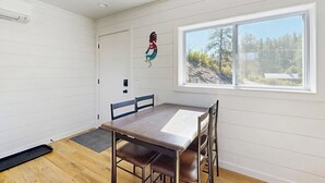 Een kamer met houten vloer, een eettafel met stoelen, een raam met uitzicht op bomen en een deur.