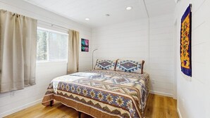 Een slaapkamer met een bed, houten vloer, witte muren, een raam met gordijnen en een kunstwerk aan de muur.