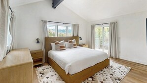 Ein Schlafzimmer mit einem großen Bett, zwei Nachttischen, einem Beistelltisch, einer Holzbank und Blick ins Freie durch die Fenster.