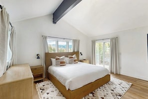 Ein Schlafzimmer mit einem großen Bett, zwei Nachttischen, einem Beistelltisch, einer Holzbank und Blick ins Freie durch die Fenster.
