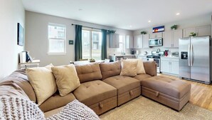 A modern living room with a sectional sofa, a kitchen area with stainless steel appliances, and a dining table.