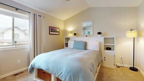 A bedroom with a bed, a ceiling fan, a window with curtains, a bedside lamp, and a small shelf with a plant.