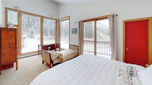 Quarto com cama, escrivaninha, cadeira, guarda-roupa e grandes janelas com vista para árvores cobertas de neve.