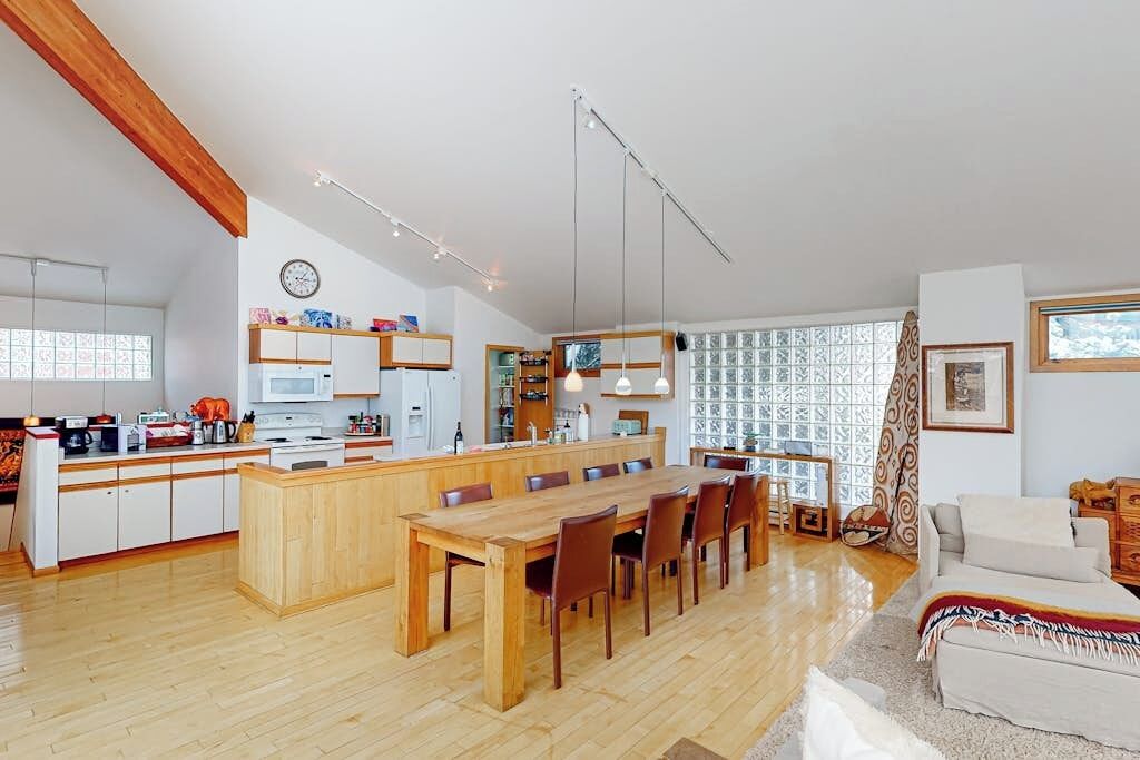 Uma sala de estar ampla com uma mesa de jantar de madeira, uma cozinha com ilha central e uma janela grande com cortinas estampadas.